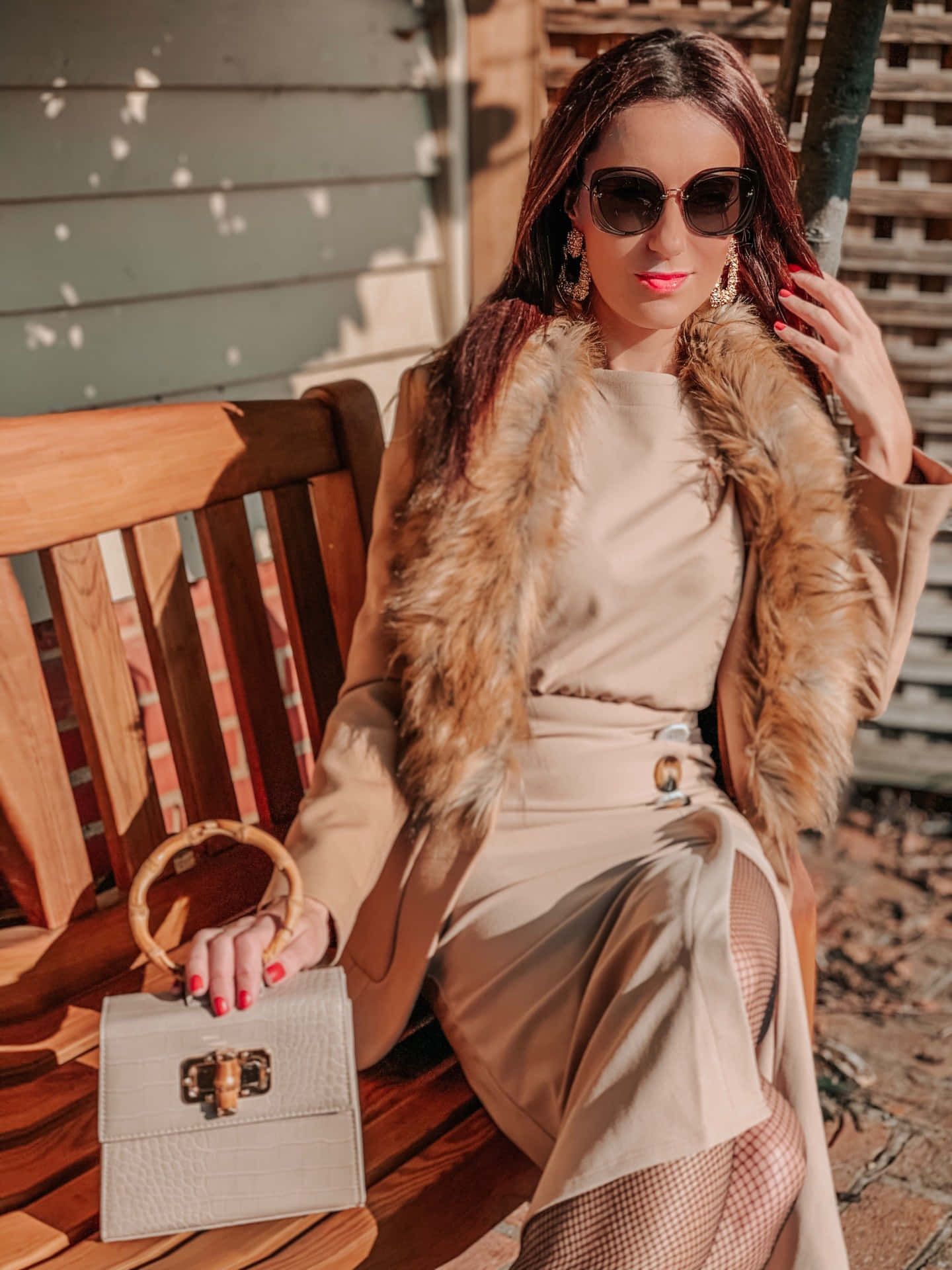 Isang babaeng naka-fur coat at beige na damit na nakaupo sa bench Wallpaper