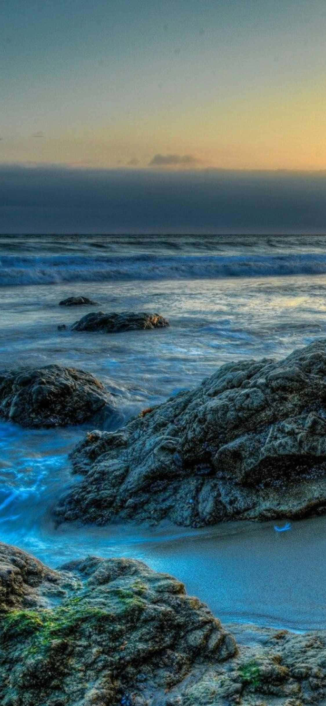 isang baybay na may mga bato at tubig sa paglubog ng araw Wallpaper