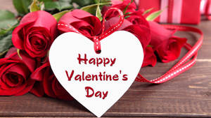 Bouquet ng pulang rosas na nakalatag sa mesa na may puting puso card na nagsasabing "Happy Valentine's Day". — Studio Display Wallpaper